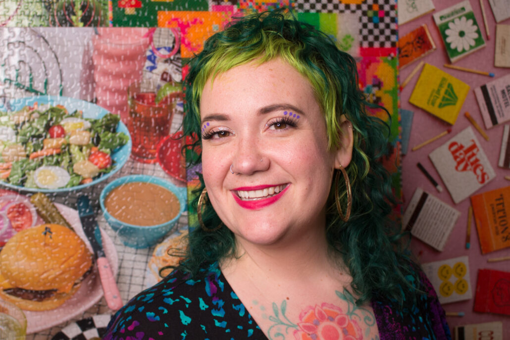 Smiling woman with green hair, standing in front of a background of puzzles
